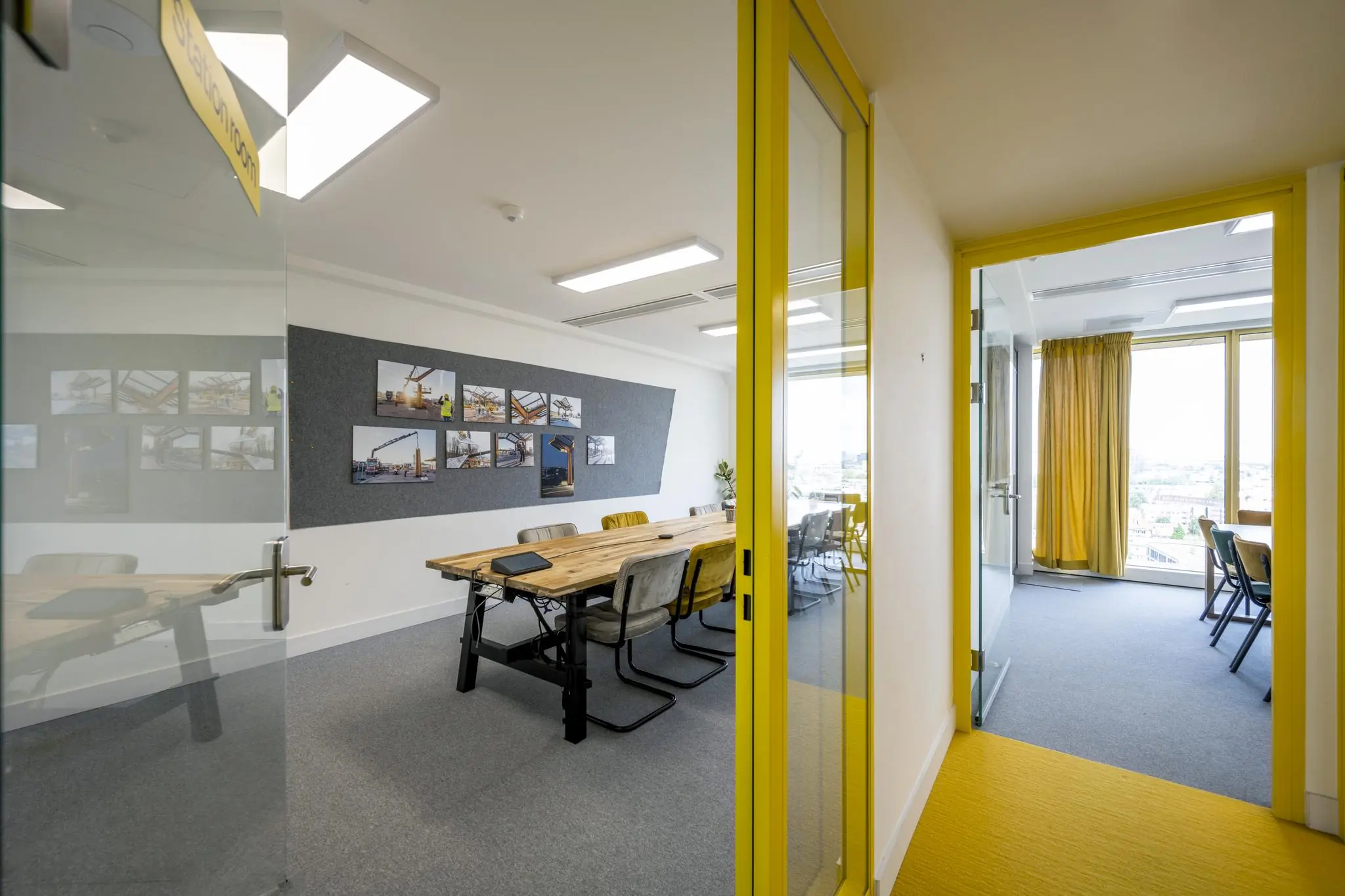 Vergaderruimte aan de James Wattstraat met lange houten tafel, gele accenten en foto's van bouwprojecten aan de muur.